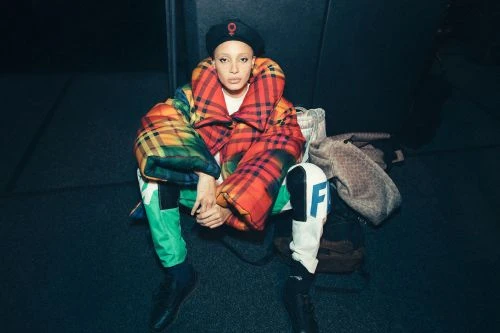 A woman with a shaved head, wearing a beret and plaid puffer jacket, is sitting on the floor with handbags in a dimly lit room.