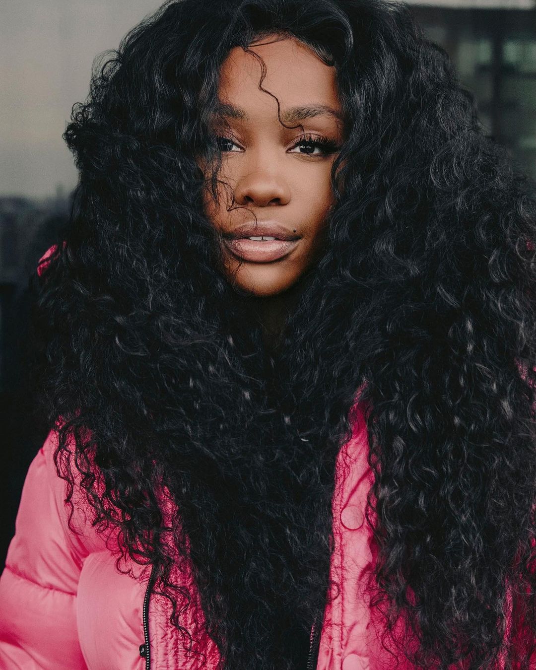 Close-up portrait of a woman with voluminous dark curly hair and a pink puffer jacket, looking directly at the camera with a neutral expression.