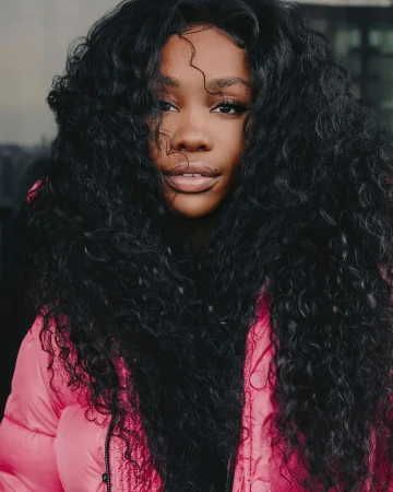 Close-up portrait of a woman with voluminous dark curly hair and a pink puffer jacket, looking directly at the camera with a neutral expression.