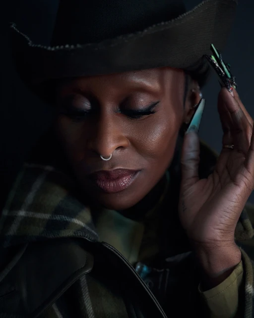 Close-up portrait of a stylish adult woman wearing a hat and jacket, eyes closed, with long decorative nails and a nose ring.
