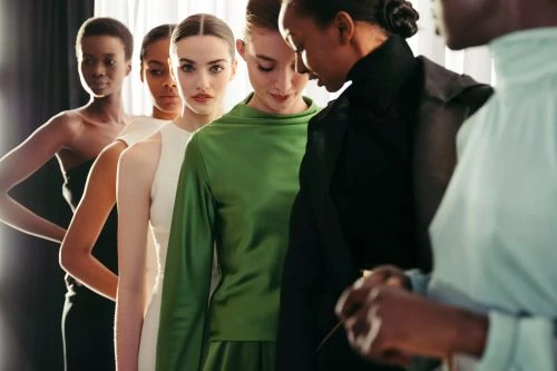 Five female models posing in a line wearing colourful dresses and high-collared tops indoors