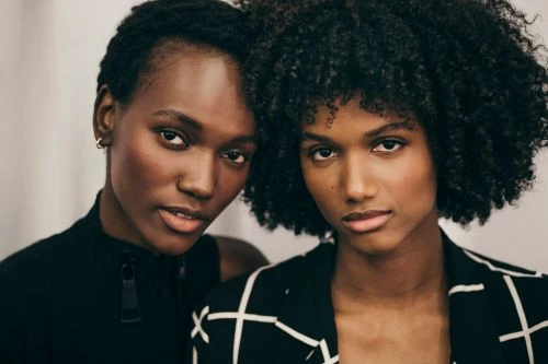 Two women of African descent pose indoors wearing fashionable clothing, showcasing beauty and style in a close-up studio shot.