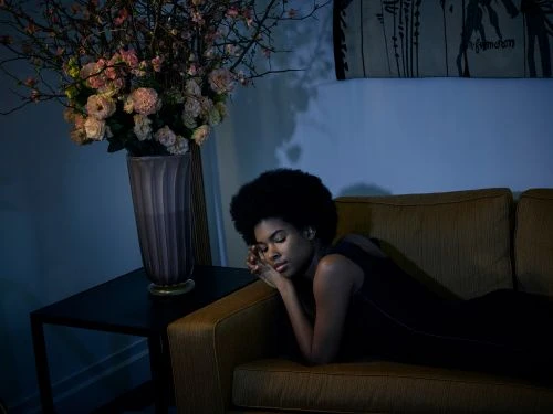 Young woman with Afro lying on a yellow couch with floral arrangement and dark interior. Relaxed pose, soft lighting, and indoor setting.