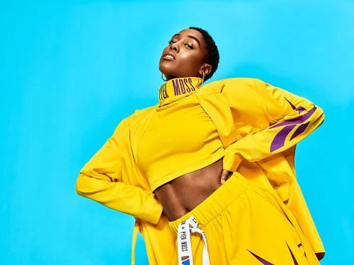 A stylish Black woman poses confidently in bright yellow athleisure wear against a vibrant turquoise backdrop. Fashion forward studio shot.