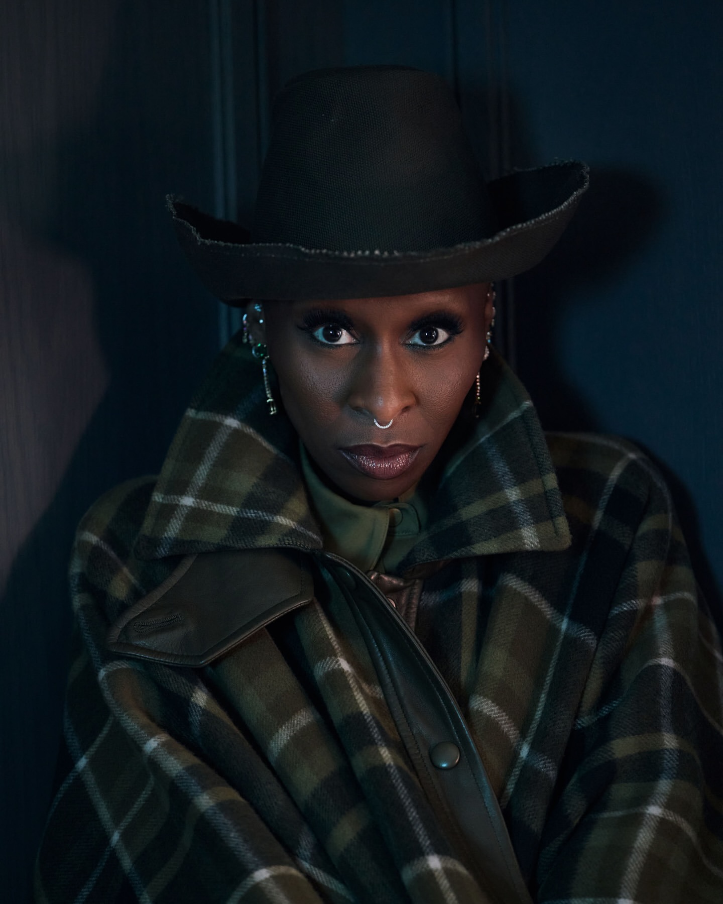 Portrait of African-descent woman wearing a hat, plaid coat, and nose ring, looking directly at the camera in a studio setting.