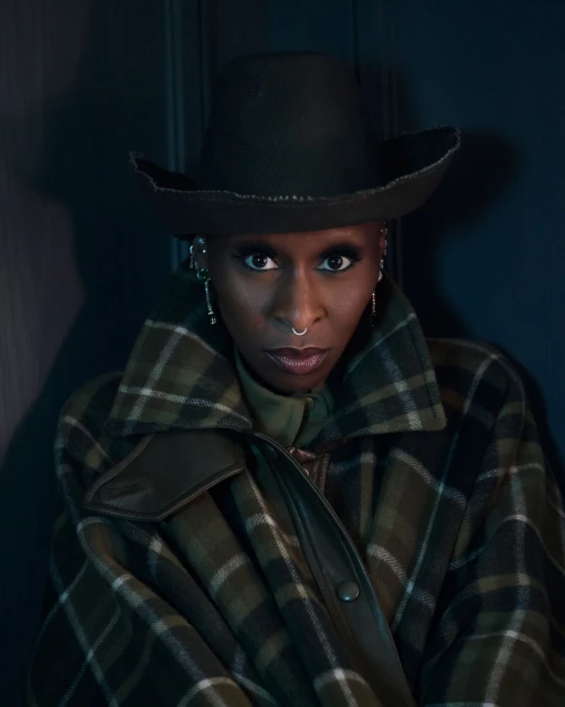 Portrait of African-descent woman wearing a hat, plaid coat, and nose ring, looking directly at the camera in a studio setting.