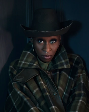 Portrait of African-descent woman wearing a hat, plaid coat, and nose ring, looking directly at the camera in a studio setting.