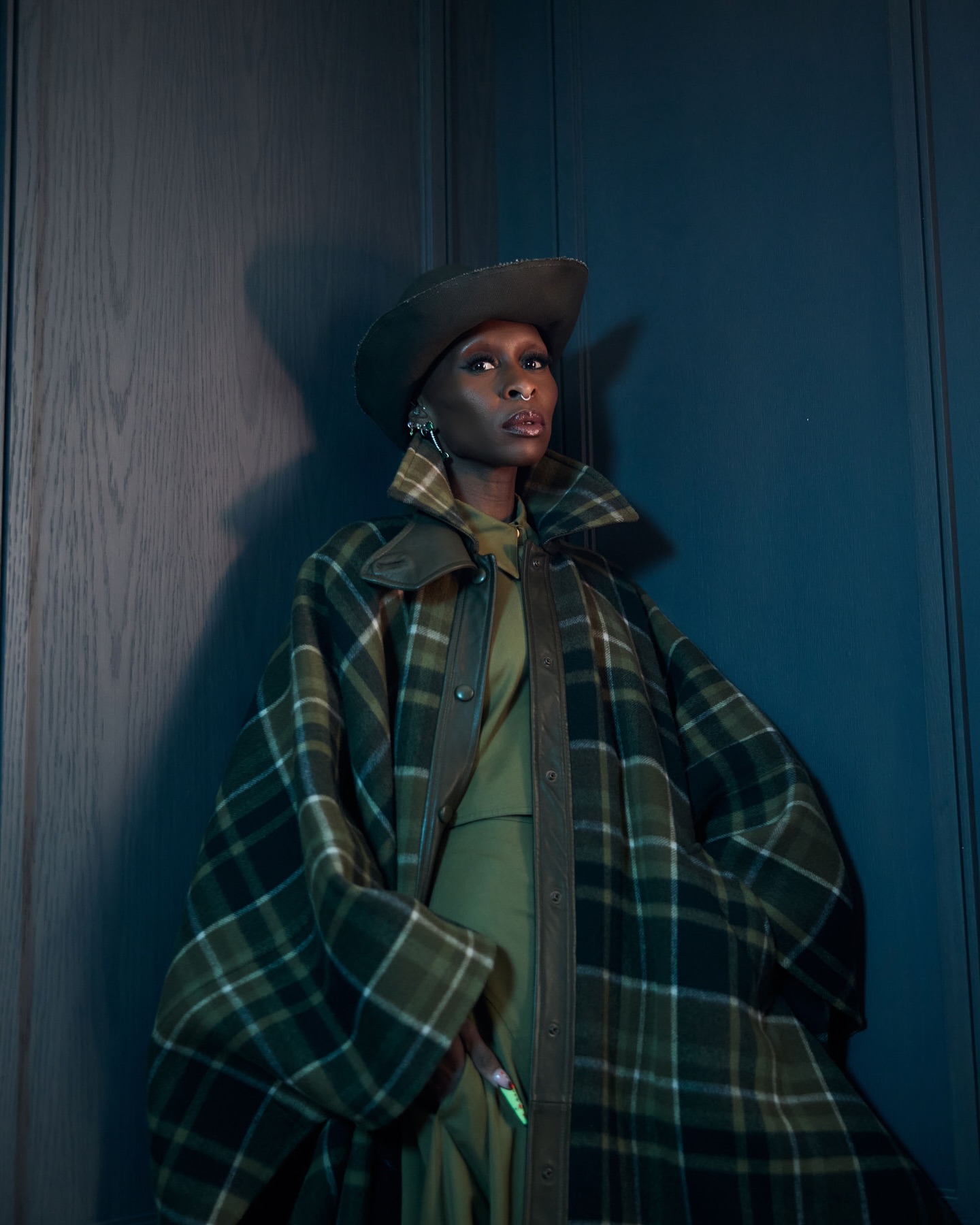 Adult woman in a green plaid coat and hat poses against a dark wall in a studio setting, looking at the camera with neutral expression.