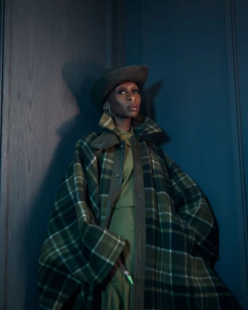 Adult woman in a green plaid coat and hat poses against a dark wall in a studio setting, looking at the camera with neutral expression.