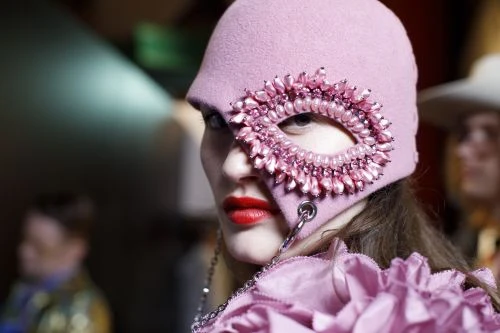 Fashionable woman wearing a pink embellished hat with red lipstick, captured in a close-up shot at eye-level in an indoor setting.