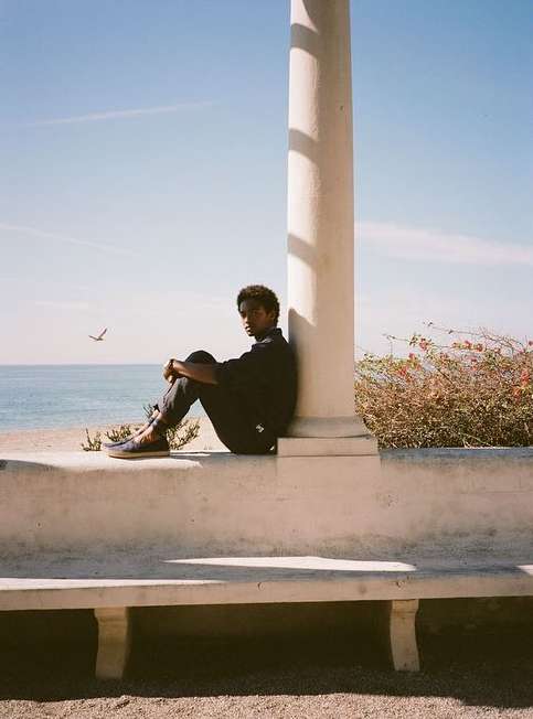 Young adult sits in front of a weathered, white column, overlooking the ocean under a clear blue sky on a sunny day outdoors.