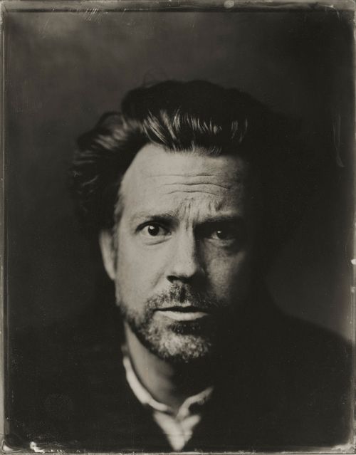 Portrait of a man with dark, wavy hair and a salt-and-pepper beard against a dark background, looking directly at the camera with a serious expression.