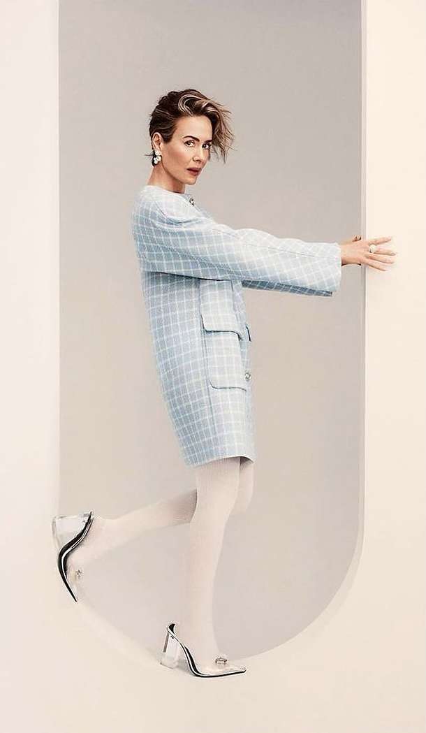 Portrait of actress Sarah Paulson wearing a light blue plaid coat and white tights posing inside an arched doorway.