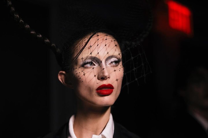 Close-up of a woman with fair skin, red lipstick, and white eye shadow wearing a black net fascinator in a dark, indoor environment.