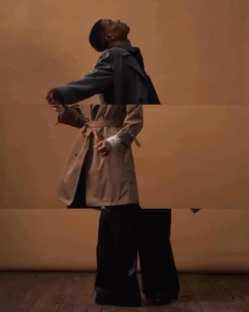 A person wearing a trench coat and wide-leg pants poses in a studio with a solid peach background and wood floor, captured with an eye-level angle.