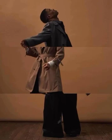 A person wearing a trench coat and wide-leg pants poses in a studio with a solid peach background and wood floor, captured with an eye-level angle.