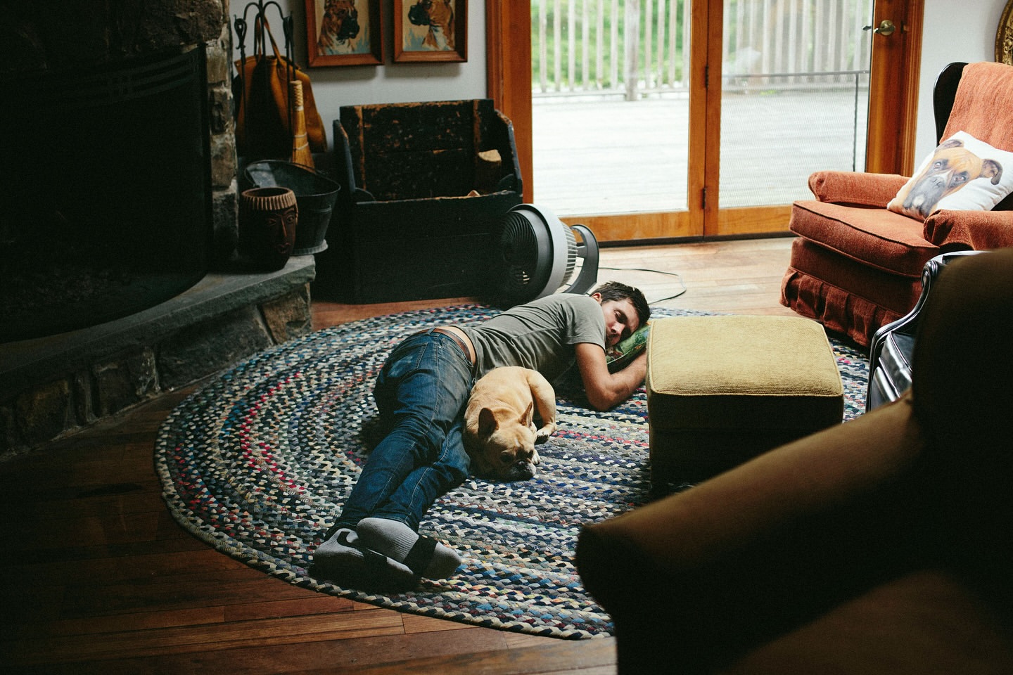 Man and dog resting together on a round braided rug indoors near a fireplace and furniture in a cozy, casual setting.