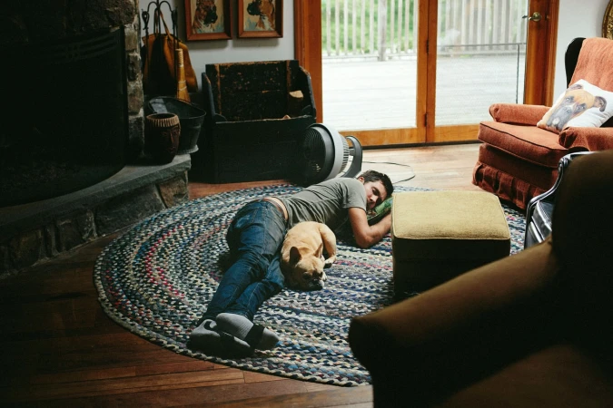 Man and dog resting together on a round braided rug indoors near a fireplace and furniture in a cozy, casual setting.