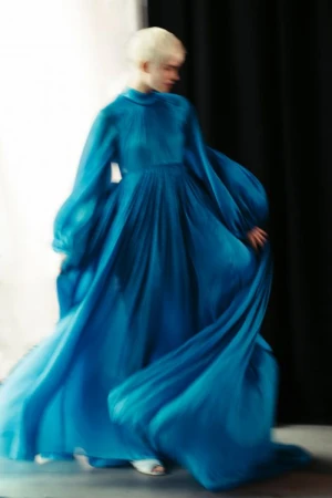 Model with platinum blonde hair wearing a flowing blue long-sleeve gown in motion against a dark curtain backdrop.