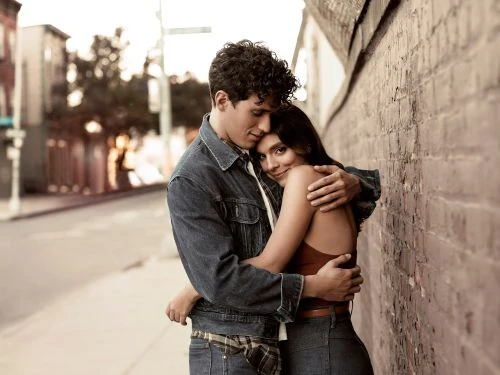 Couple embraces on city sidewalk next to brick wall on a sunny day; man in denim jacket, woman in brown tank top and denim jeans.