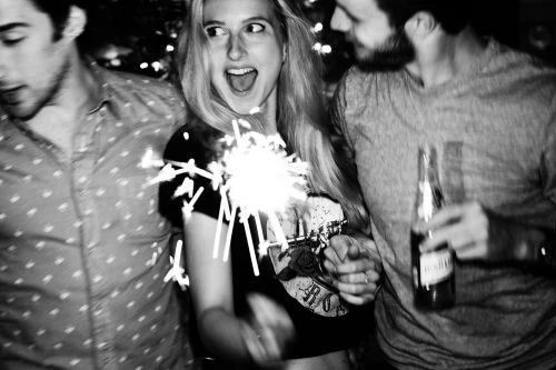 Three people celebrate with a sparkler and a beer bottle in a grainy, black-and-white, low-angle shot. A woman smiles, holding the sparkler.