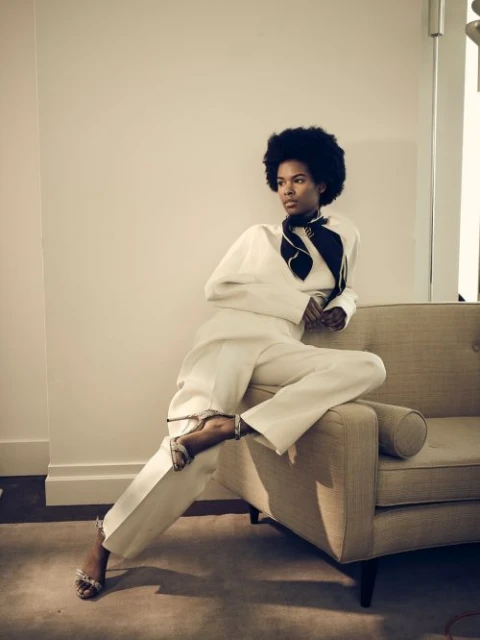 An elegant woman with dark curly hair wearing a white suit, black scarf, and silver high heels sitting on a tan mid-century modern chair.