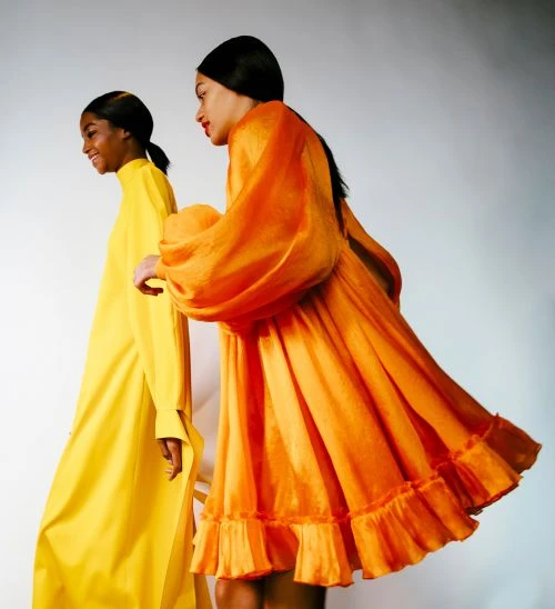 Two Black women wearing bright dresses, one yellow and one orange, stand against a white background in a fashion forward studio shot.