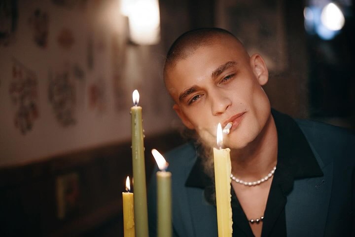 Portrait of a young adult male smoking a cigarette, with multiple lit candles and a dark background, in a shallow depth of field.