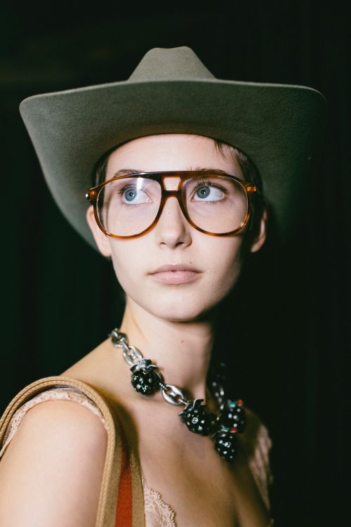 Close-up portrait of a woman wearing large aviator glasses, an olive cowboy hat, and a chunky silver necklace by Victoria Will.
