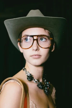 Close-up portrait of a woman wearing large aviator glasses, an olive cowboy hat, and a chunky silver necklace by Victoria Will.