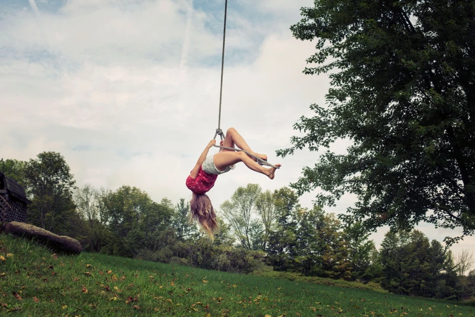 A blonde woman in a red shirt hangs upside down on a rope swing, with a grassy hill, trees, and cloudy sky in the background.