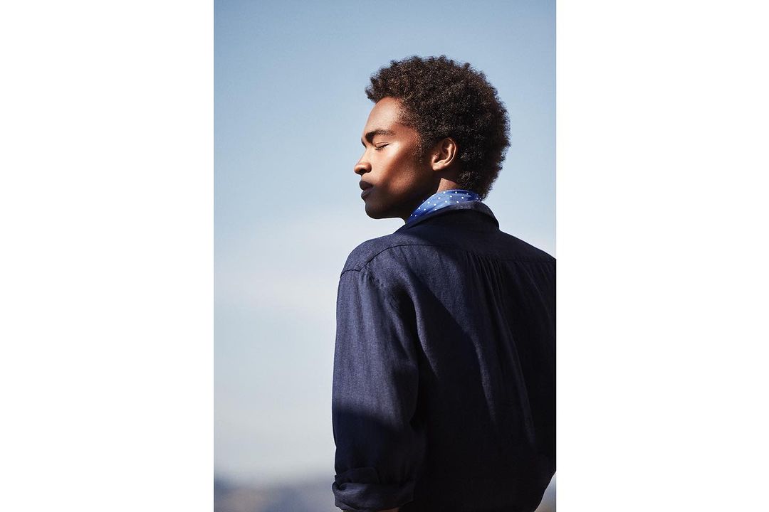 Profile of an adult male with afro hairstyle wearing a blue button-down shirt and a patterned neckerchief against a clear, light-blue sky.