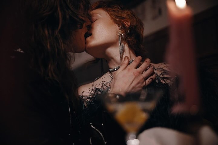 Close-up of a romantic couple kissing indoors with a cocktail glass and lit candle in the foreground, creating a moody atmosphere.