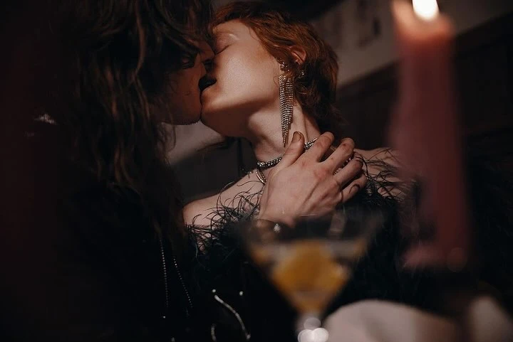 Close-up of a romantic couple kissing indoors with a cocktail glass and lit candle in the foreground, creating a moody atmosphere.