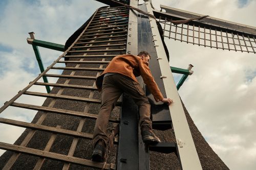 Adult male ascends a wooden ladder attached to a historic windmill, showcasing architectural detail against a cloudy sky backdrop.