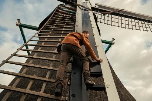 Adult male ascends a wooden ladder attached to a historic windmill, showcasing architectural detail against a cloudy sky backdrop.