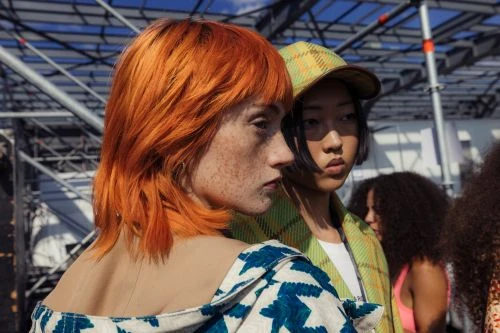 Two models with unique hairstyles stand side by side in a candid outdoor setting, showcasing distinct fashion and beauty.