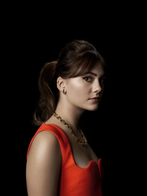 Portrait of a woman with dark hair in a ponytail wearing a red dress and gold jewelry against a black backdrop in a photo studio.