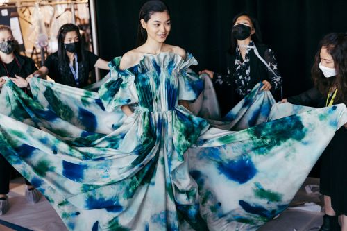 A young Asian model in a blue and green dress is helped by a group of women backstage at a fashion show, photo studio environment.