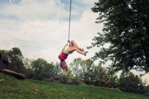 Woman upside down on rope swing over a grassy hill, surrounded by green trees, with a cloudy sky in the background.