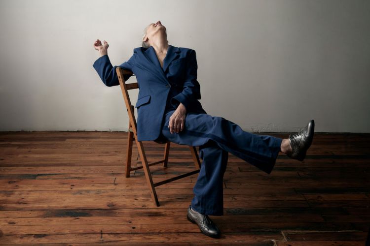 Woman in a blue suit sitting backwards on a wooden chair with a white wall and wood floor in the background, looking up.