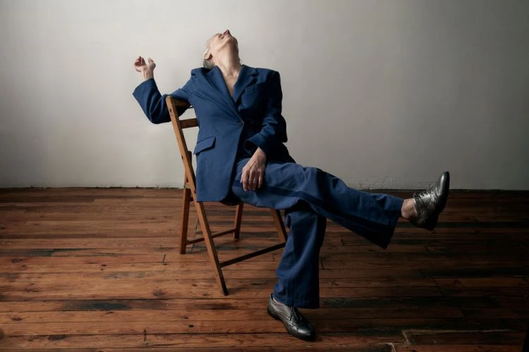 Woman in a blue suit sitting backwards on a wooden chair with a white wall and wood floor in the background, looking up.