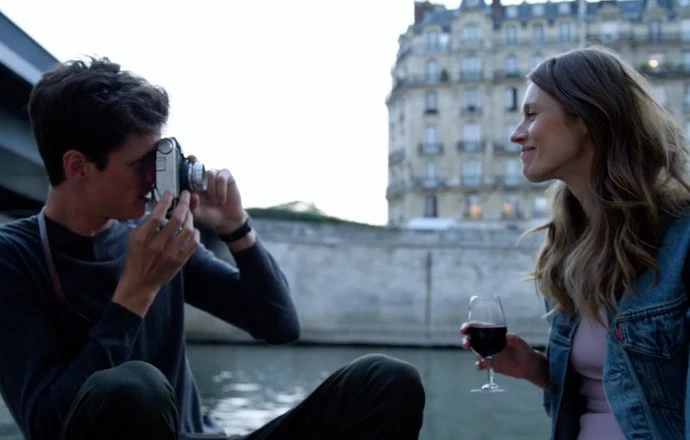 A young couple in Paris, France. The man takes a photo of the woman with a vintage camera. She holds a wine glass with a smile.