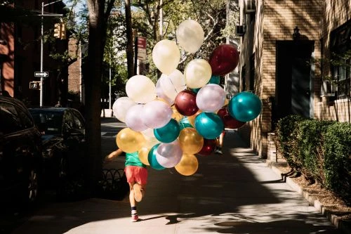 Young person runs on a city sidewalk holding a large bunch of colorful helium balloons, with brick buildings and trees in the background.