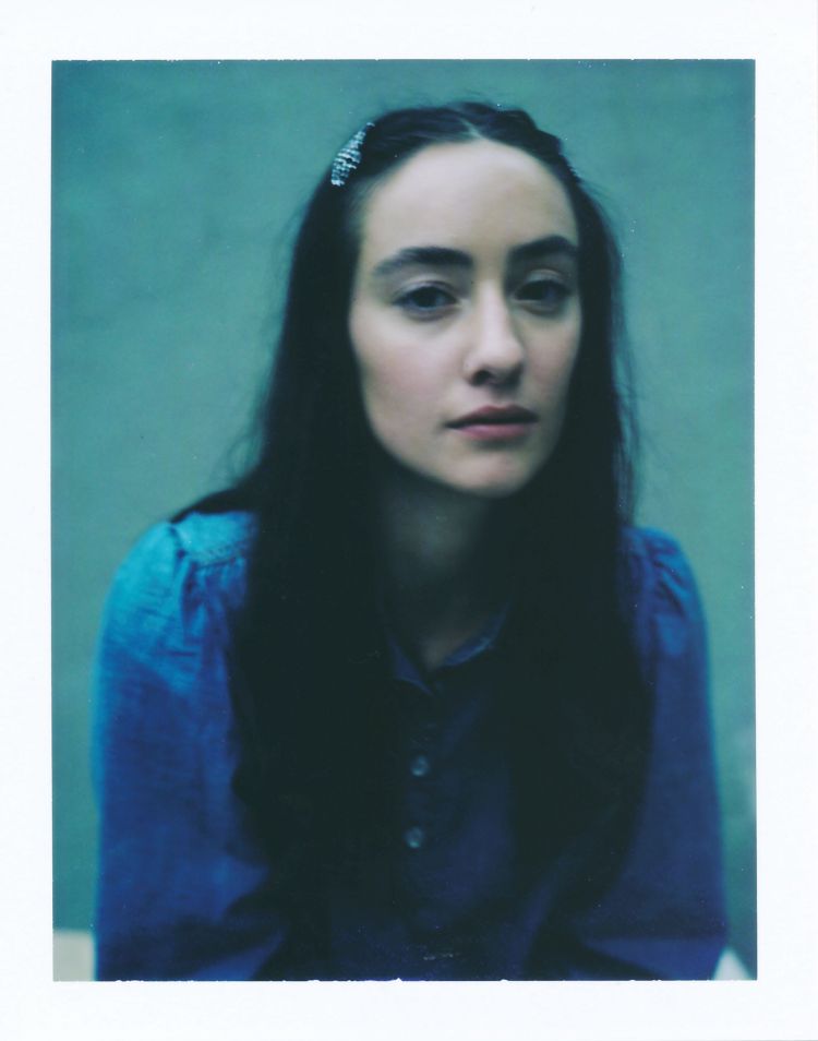 Close-up portrait of a woman with dark hair, wearing a blue blouse, against an aquamarine background. Medium studio shot with soft lighting.