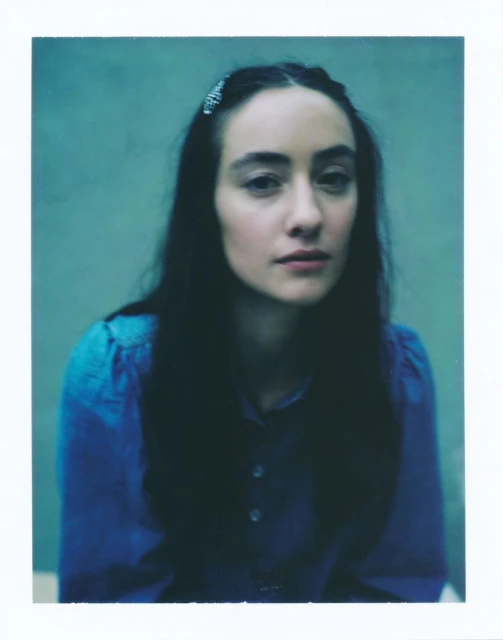 Close-up portrait of a woman with dark hair, wearing a blue blouse, against an aquamarine background. Medium studio shot with soft lighting.
