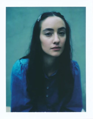 Close-up portrait of a woman with dark hair, wearing a blue blouse, against an aquamarine background. Medium studio shot with soft lighting.
