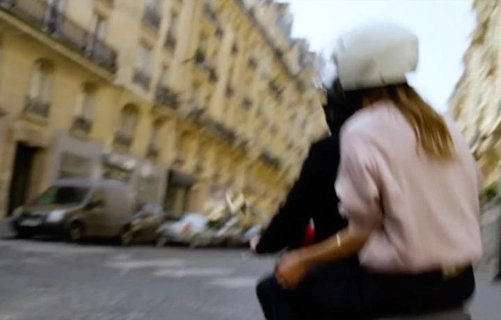 Two people ride a scooter through European city streets, passing parked cars and sandstone buildings on a sunny day.