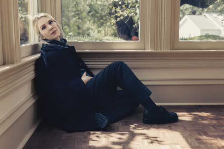 Woman in dark coat seated on parquet flooring leaning against a window frame by Victoria Will