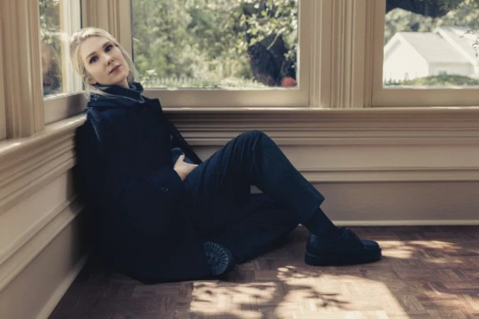 Woman in dark coat seated on parquet flooring leaning against a window frame by Victoria Will